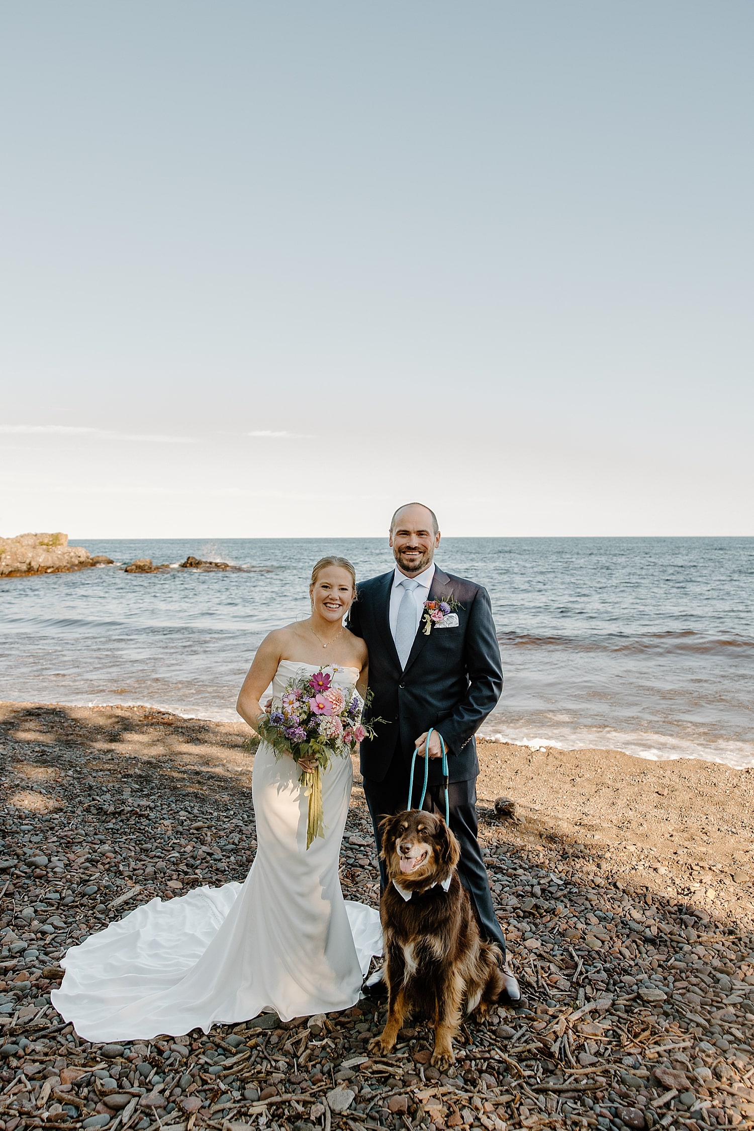 newlyweds stand near the waters edge with pup by Samantha Burke Photography