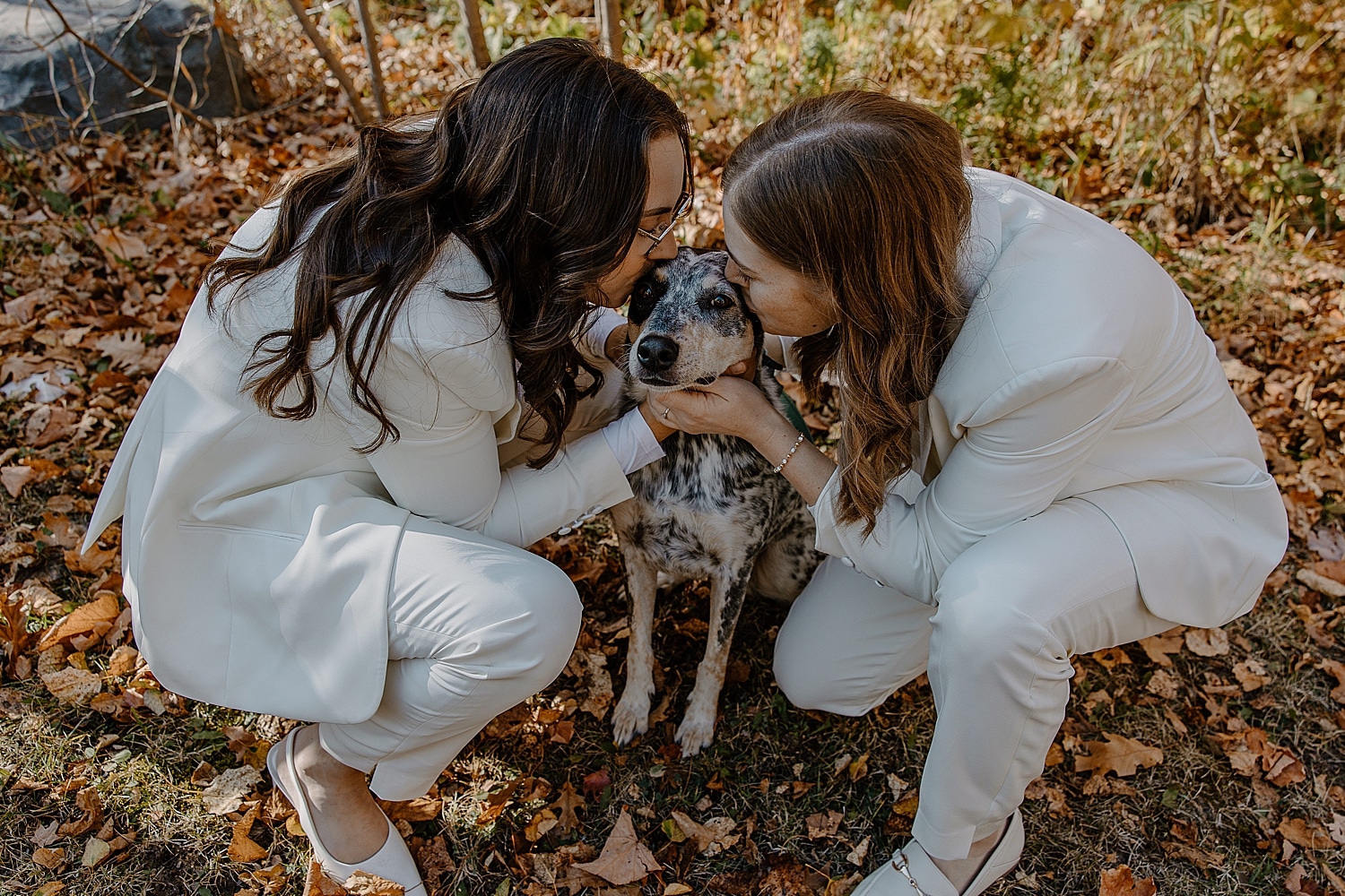 two women in white suits kiss spotted dog by Wisconsin wedding photographer