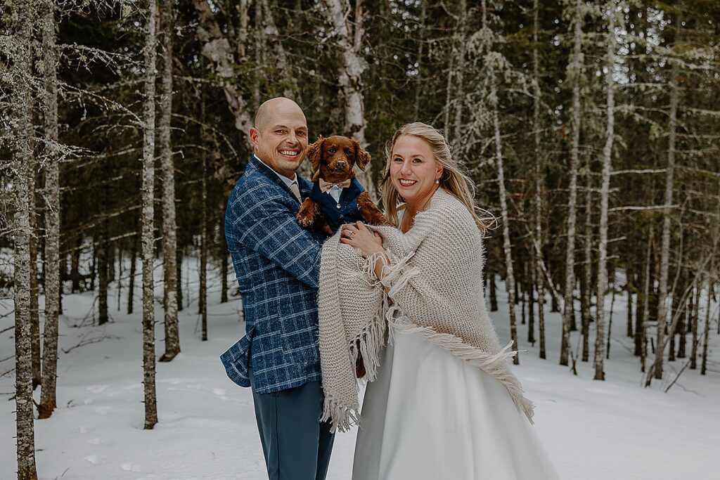 winter bride and groom stand in the trees holding their pet by Wisconsin wedding photographer